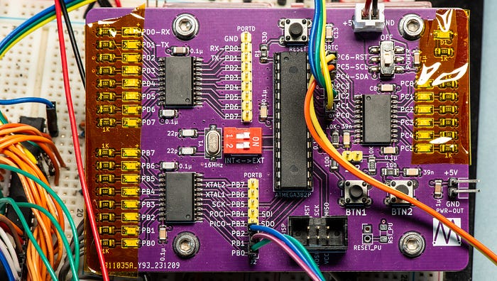 A close-up photograph of an electronic circuit board to which are connected multiple wires. Some components at the left and right edges are covered in transparent yellow tape.