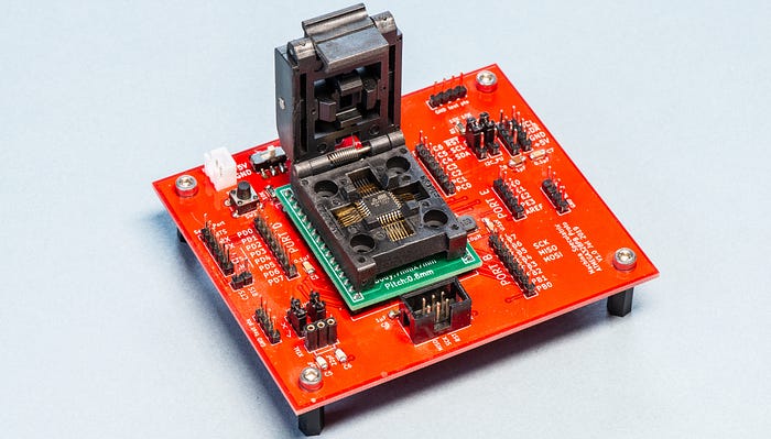 Photograph of a red electronics circuit board in the centre of which is a large, black socket. The cover of the socket is flipped open to reveal a small chip inside.