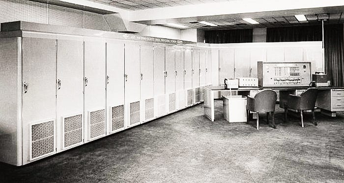 1950s black and white photograph of a mainframe computer showing multiple processor cabinets and a desk-based control station. The computer essentially fills a large room.