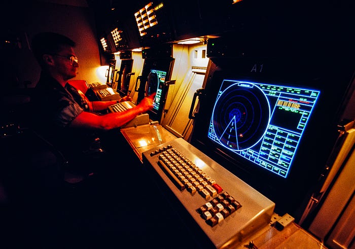 Photograph of a row of computer screens and keyboards in a dimly lit room with a barely seen person on the left.
