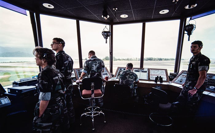 Photograph of the inside of an air traffic control tower at an airport with five people, four of them standing, looking out of the windows. All five people are in military camouflage uniforms.