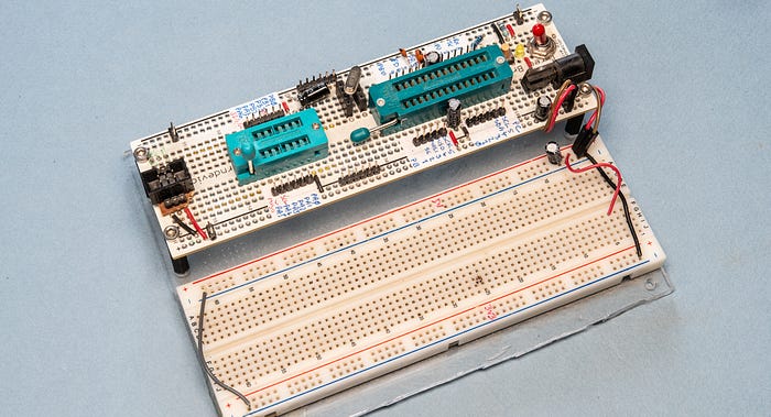 Photograph of an electronic circuit created on prototyping board, including two turquoise chip sockets. Alongside it is a breadboard and both boards are mounted on an acrylic base.