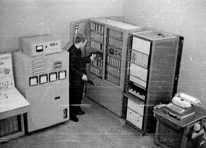 Black and white photograph of a man standing amidst the cabinets of a 1960s-era computer.