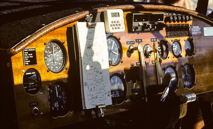 Photograph of a wooded instrument panel in an old aircraft, lit by a low sun. A notepad with strange handwritten symbols is clipped to the panel.